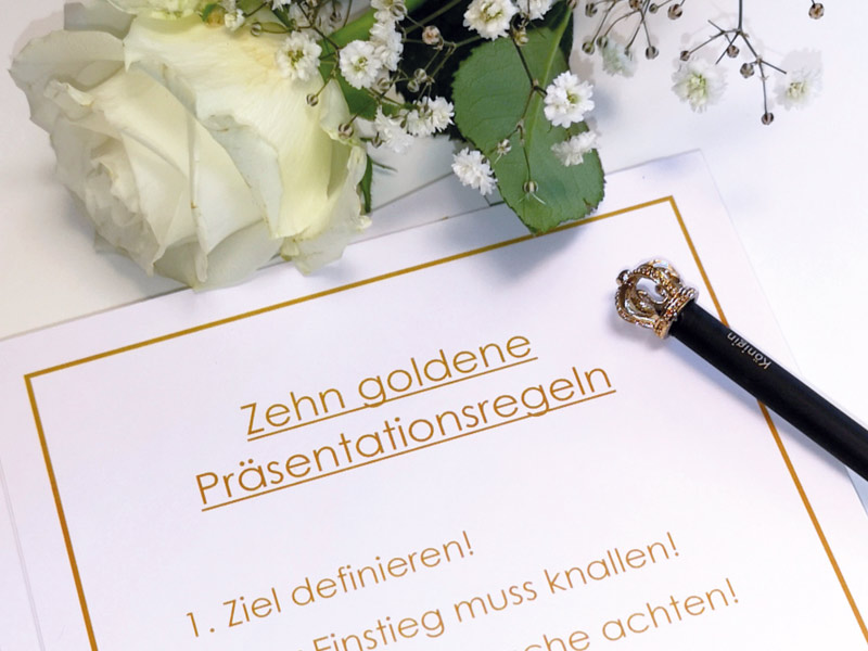 Auf dem Foto sind die 10 goldenen Präsentationsregeln zu sehen, eine weiße Rose und ein Bleistift mit einer Krone.